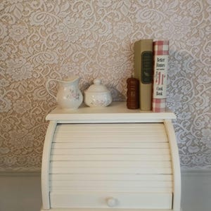May include: A white, roll-top breadbox with a small drawer, set on a wooden stand. A shelf above holds a small pitcher and sugar bowl with floral designs, a pepper grinder, and two cookbooks. The background features a lace-patterned wallpaper.
