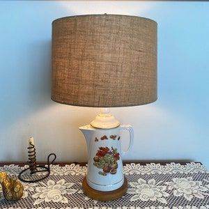 May include: A white ceramic lamp shaped like a coffee pot with a brown burlap lampshade. The coffee pot has a design of grapes and nuts on it. The lamp is sitting on a wooden base and a white lace table runner.