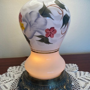 May include: White ceramic table lamp with a floral design featuring purple, pink, yellow, and green flowers. The lamp has a gold trim and a brass base with ornate details.