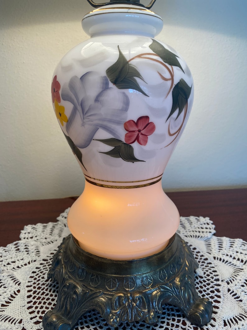 May include: White ceramic table lamp with a floral design featuring purple, pink, yellow, and green flowers. The lamp has a gold trim and a brass base with ornate details.