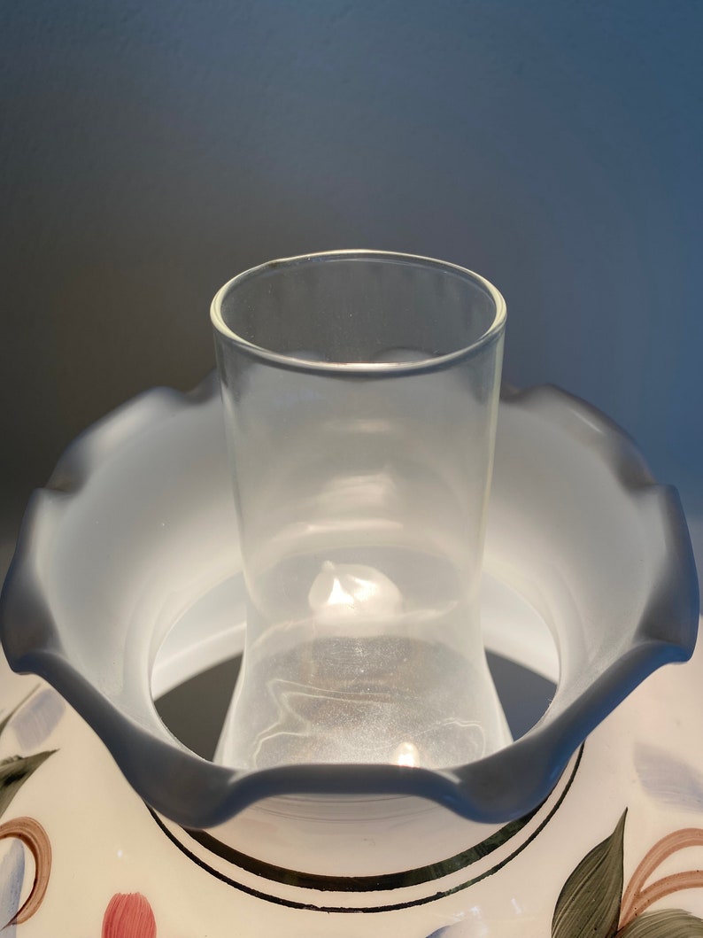 May include: A white ceramic lampshade with a scalloped edge and a floral design. The lampshade has a clear glass chimney inside.