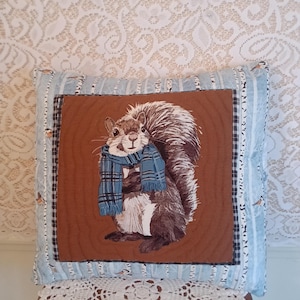 May include: A decorative square pillow featuring a squirrel wearing a blue scarf. The squirrel is centered on a brown background with a wood grain pattern. The pillow has a blue and white striped border with small bird accents.