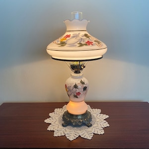 May include: A white table lamp with a floral design on the base and shade. The lamp is turned on and the light is shining through the white glass.