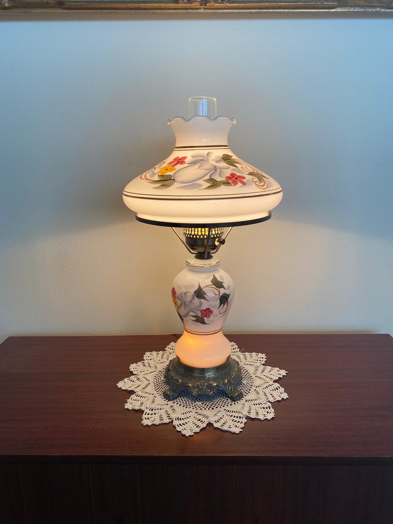May include: A white table lamp with a floral design on the base and shade. The lamp is turned on and the light is shining through the white glass.