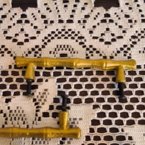 May include: Two gold-colored cabinet pulls with a textured bamboo-inspired design. Each handle features two vertical mounting posts with black threaded screws extending from the base, designed for attachment to furniture or cabinetry doors.