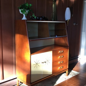 May include: A mid-century modern cabinet with a wood frame and white doors. The doors have a starburst design and gold-colored hardware. The cabinet has glass shelves and drawers.