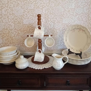 May include: A collection of vintage cream-colored dishware with a floral pattern. Includes plates, bowls, cups, a creamer, and a sugar bowl with a lid. A wooden cup rack holds several cups. The plates are stacked on a black stand.