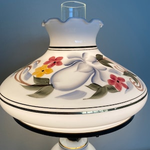 May include: White ceramic table lamp with a floral design. The lampshade has a scalloped edge and features painted flowers in shades of red, yellow, and blue.