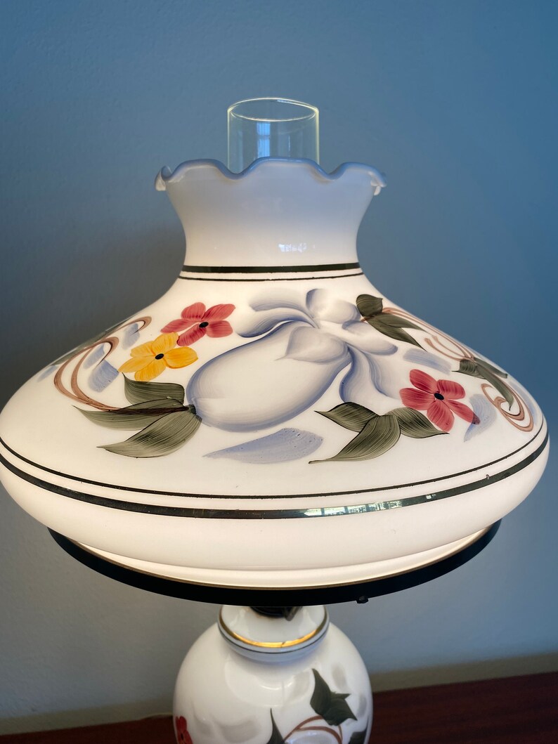 May include: White ceramic table lamp with a floral design. The lampshade has a scalloped edge and features painted flowers in shades of red, yellow, and blue.