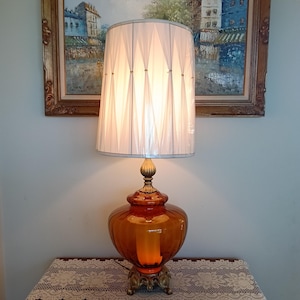 May include: A table lamp featuring a large, amber-colored glass base with a ribbed texture and a gold-toned metal pedestal. The lamp is topped with a white, pleated fabric drum shade that creates a diamond pattern effect when illuminated.