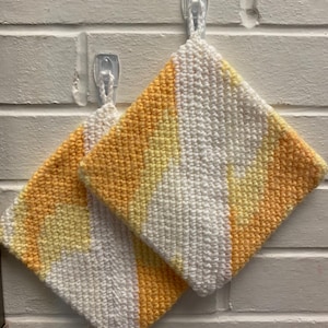 May include: Two square, handmade potholders with a loop for hanging. The potholders are crocheted with a pattern of white, yellow, and orange stripes. They are hanging on clear adhesive hooks against a white brick wall.
