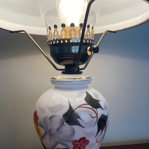 May include: A white ceramic lamp with a floral design. The lamp has a gold trim and a clear glass shade with a brass finial.
