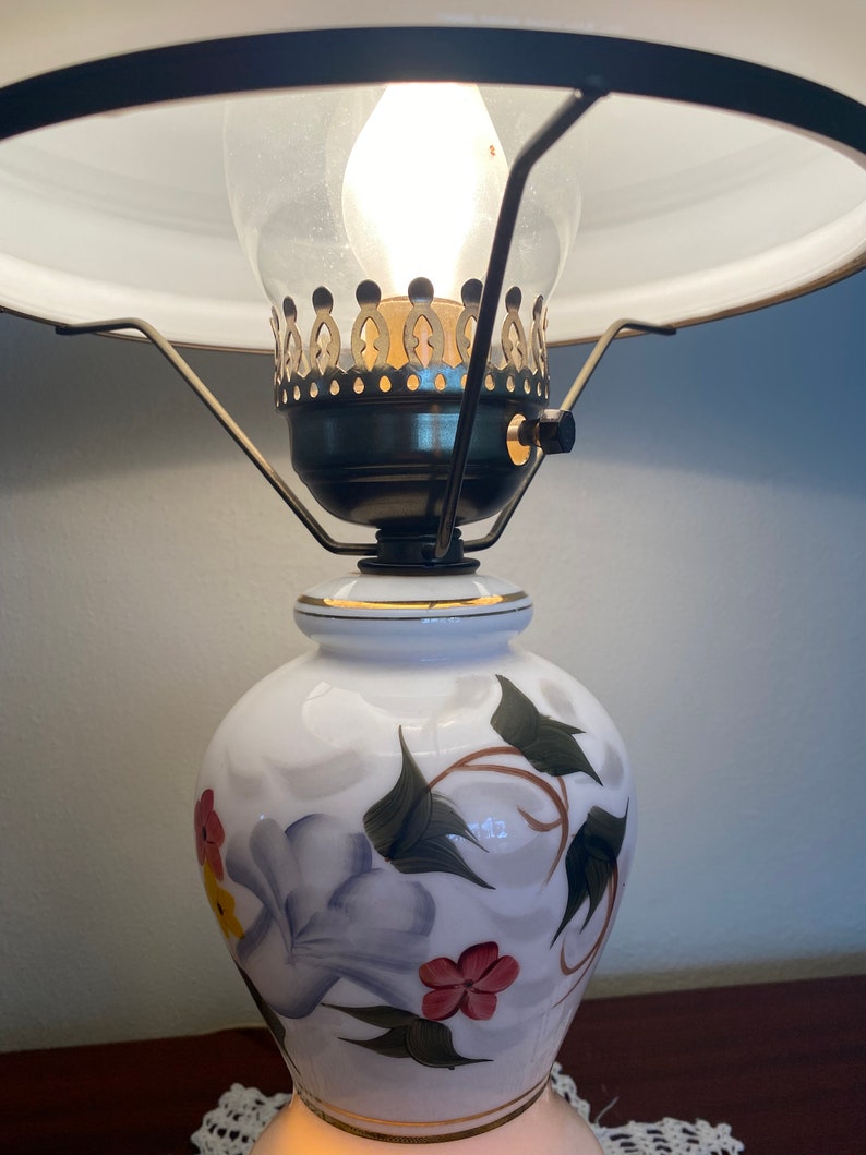 May include: A white ceramic lamp with a floral design. The lamp has a gold trim and a clear glass shade with a brass finial.