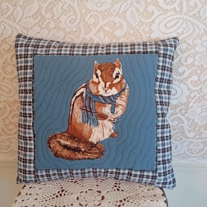 May include: Square decorative pillow featuring a chipmunk illustration. The chipmunk is wearing a blue and white plaid scarf and is set against a blue background with a quilted texture. The pillow has a plaid border in shades of blue, black, and white.