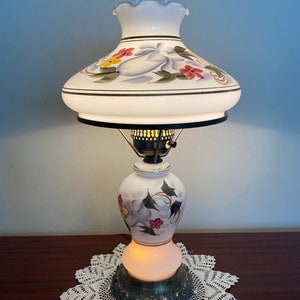 May include: A white ceramic table lamp with a floral design. The lamp has a white shade with a scalloped edge and a gold trim. The base of the lamp is a white ceramic vase with a floral design. The lamp is sitting on a white doily.