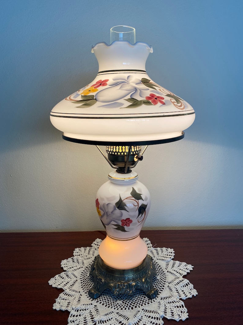 May include: A white ceramic table lamp with a floral design. The lamp has a white shade with a scalloped edge and a gold trim. The base of the lamp is a white ceramic vase with a floral design. The lamp is sitting on a white doily.
