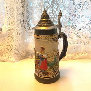 May include: A ceramic beer stein with a lid and handle. The stein features a scene of people in traditional clothing in a mountainous landscape.