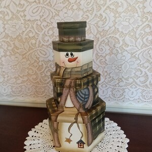 May include: A stack of three hexagonal boxes painted to resemble a snowman. The top box is a dark green hat, the middle box features a snowman face with an orange carrot nose, and the bottom box displays a plaid scarf and small star ornaments.