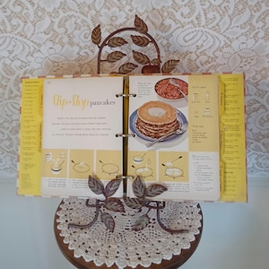 May include: An open cookbook displays a recipe for "Flip-flop pancakes." The vintage cookbook is held by a decorative metal stand with leaf accents. The recipe includes illustrations and ingredients for the pancakes and a cranberry-orange honey sauce.