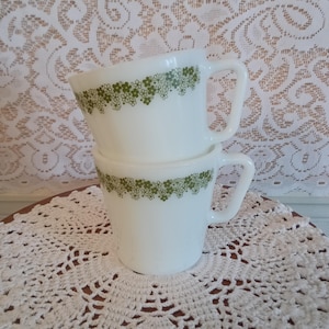 May include: Two white ceramic mugs stacked on top of each other. Each mug has a handle and is decorated with a band of small green floral designs around the rim. The mugs are on a white crocheted doily.