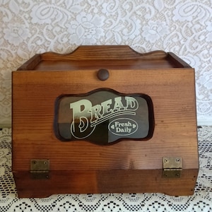 May include: A medium-brown wooden bread box with a hinged front door featuring a central glass window. The glass displays the text BREAD in large letters above the smaller text Fresh Daily. The door has a small round knob and two brass hinges.