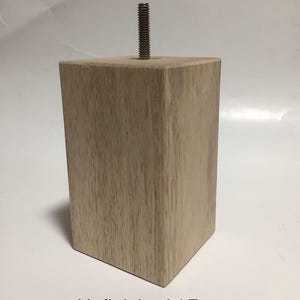 May include: Unfinished wooden furniture leg with a threaded metal insert. The leg is square and has a light brown color. The text "Unfinished / Raw" is visible below the leg.