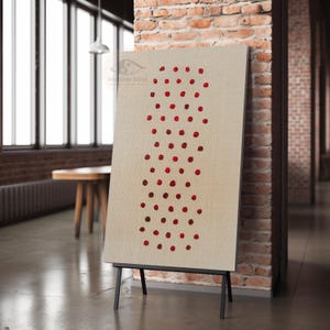 May include: A beige canvas art piece with a vertical pattern of red dots, displayed on a black easel. The artwork is set against a backdrop of a brick wall and a window, creating a modern, minimalist aesthetic.