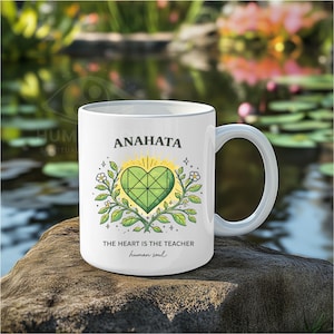 Könnte beinhalten: Weißer Keramikbecher mit dem Wort "ANAHATA" und einem grünen Herzdesign in der Mitte, umgeben von Blättern und Blumen. Darunter steht der Text "THE HEART IS THE TEACHER human soul". Der Becher hat einen gebogenen Griff.