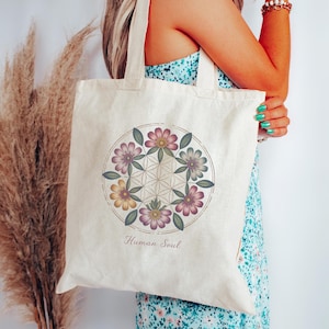 May include: Cream-colored tote bag featuring a floral design within a circular frame. The design includes pink, purple, and yellow flowers with green leaves, and the words "Human Soul" are printed below the design. The bag has long handles.