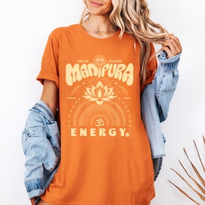 Könnte beinhalten: Orangefarbenes T-Shirt mit dem Wort "Manipura" in Retro-Schrift, einer Lotusblume und dem Wort "Energy". Das Shirt zeigt auch die Wörter "Solar Plexus", "Human" und "Soul".