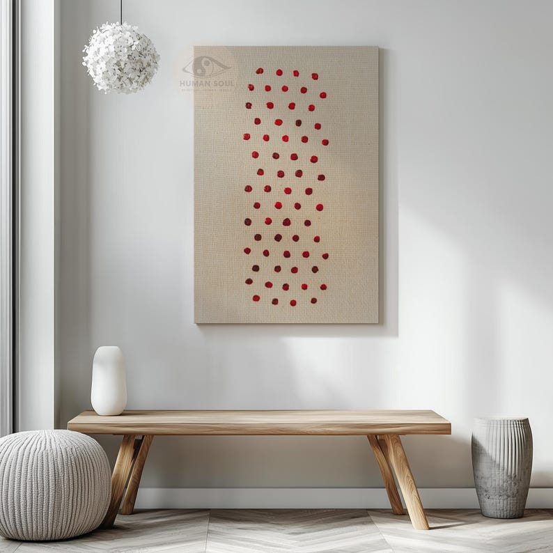 May include: A rectangular canvas art piece with a beige background and a vertical pattern of red dots. The artwork hangs on a white wall above a wooden bench. A white vase and a gray ottoman are in the foreground.