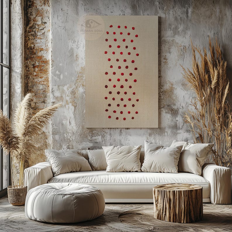 May include: A beige canvas wall art piece with a pattern of red dots. The artwork hangs above a white sofa with several pillows. A round ottoman and a wooden stump table are in front of the sofa.