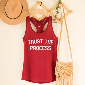 Op de afbeelding: Een rode tanktop met de witte tekst "TRUST THE PROCESS". Een gouden ketting hangt aan de hanger. Een beige hoed en een macramé tas zijn ook zichtbaar.