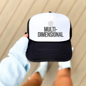 May include: A black and white trucker hat with the text "MULTI-DIMENSIONAL" in a bold, sans-serif font. The hat features a white front panel and a black brim and mesh back. A geometric design is above the text.
