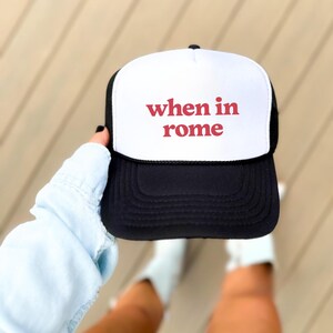 May include: A black and white trucker hat featuring the text "when in rome" in red. The hat has a white front panel, a black peak, and a black mesh back. A fashionable accessory for everyday wear.