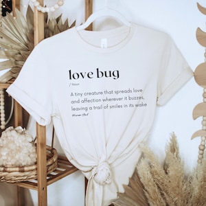 May include: A beige t-shirt with a black graphic that reads "love bug / Noun A tiny creature that spreads love and affection wherever it buzzes, leaving a trail of smiles in its wake How Gud"