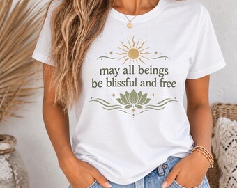 Può includere: Una t-shirt bianca a maniche corte con una stampa grafica raffigurante un sole dorato, un fiore di loto verde e il testo "may all beings be blissful and free" in lettere verde scuro, centrato sul petto del capo.