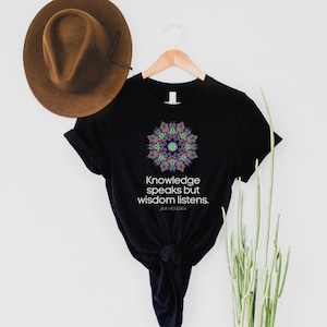 Op de afbeelding: Zwart T-shirt met een kleurrijk mandala-ontwerp en de quote "Knowledge speaks but wisdom listens." van Jimi Hendrix.