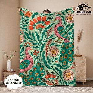 May include: A plush blanket featuring a repeating design of pink peacocks, flowers, and foliage in green, orange, and pink hues. The blanket has the words "PLUSH BLANKET" in a white box. The blanket is held up by a person.