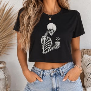 Könnte beinhalten: Ein schwarzes, kurzärmeliges bauchfreies Oberteil mit einer weißen grafischen Illustration eines menschlichen Skeletts, das eine kleine Topfpflanze hält. Das Shirt hat einen Rundhalsausschnitt und einen geraden Saum.