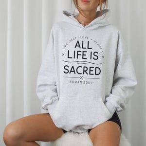 Puede incluir: Una sudadera con capucha gris claro con un bolsillo delantero y una capucha con cordón. La sudadera con capucha presenta el texto "ALL LIFE IS SACRED" en negro, rodeado por las palabras "RESPECT, LOVE, PEACE" en un diseño circular. La frase "HUMAN SOUL" también está impresa en la sudadera.