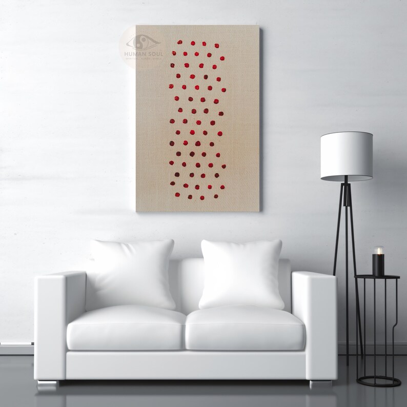 May include: A rectangular canvas art piece with a beige background features a vertical pattern of red dots. The artwork hangs above a white sofa with two pillows. A black floor lamp and side table with a candle are also visible.