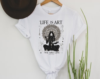 Op de afbeelding: Wit T-shirt met een zwart-wit grafisch ontwerp met een vrouw die zit met een penseel en een palet. De tekst "Life is Art - We are Life" staat op het shirt gedrukt.