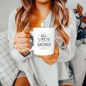May include: White ceramic mug with the text "ALL LIFE IS SACRED" in black, along with the words "Respect, Love, Peace" and "Human Soul". The mug has a curved handle and is held by a person.