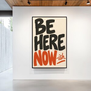 Op de afbeelding: Een rechthoekig wandkunstwerk met een crèmekleurige achtergrond en handgeschilderde letters. De woorden BE HERE zijn in het zwart, terwijl het woord NOW-ish in fel oranje is weergegeven, alles gevat in een dunne zwarte lijst.
