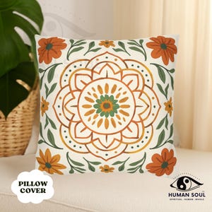 May include: Square pillow cover with a cream background featuring a central mandala design in shades of orange, yellow, and green. The design is surrounded by orange flowers and green leaves. The words "PILLOW COVER" are printed on the bottom left.