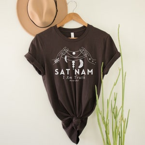 Puede incluir: Una camiseta marrón oscuro con un diseño gráfico blanco que representa una mano estilizada sosteniendo una cadena de cuentas y plumas. El texto "SAT NAM I Am Truth" está impreso debajo del gráfico.
