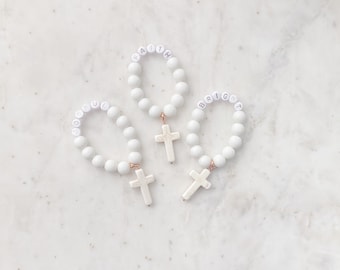 May include: Three white beaded bracelets with a white cross charm. Each bracelet has a different name spelled out on it: "Faith", "Joyful", and "Bright".