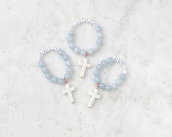 May include: Three light blue beaded bracelets with white cross pendants. Each bracelet has a different word spelled out in white letter beads: "JOYFUL", "BLESSED", and "ADORE". The bracelets are displayed on a white marble surface.
