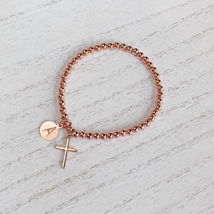 May include: A rose gold beaded bracelet with a small cross charm and a round charm engraved with the letter "A".